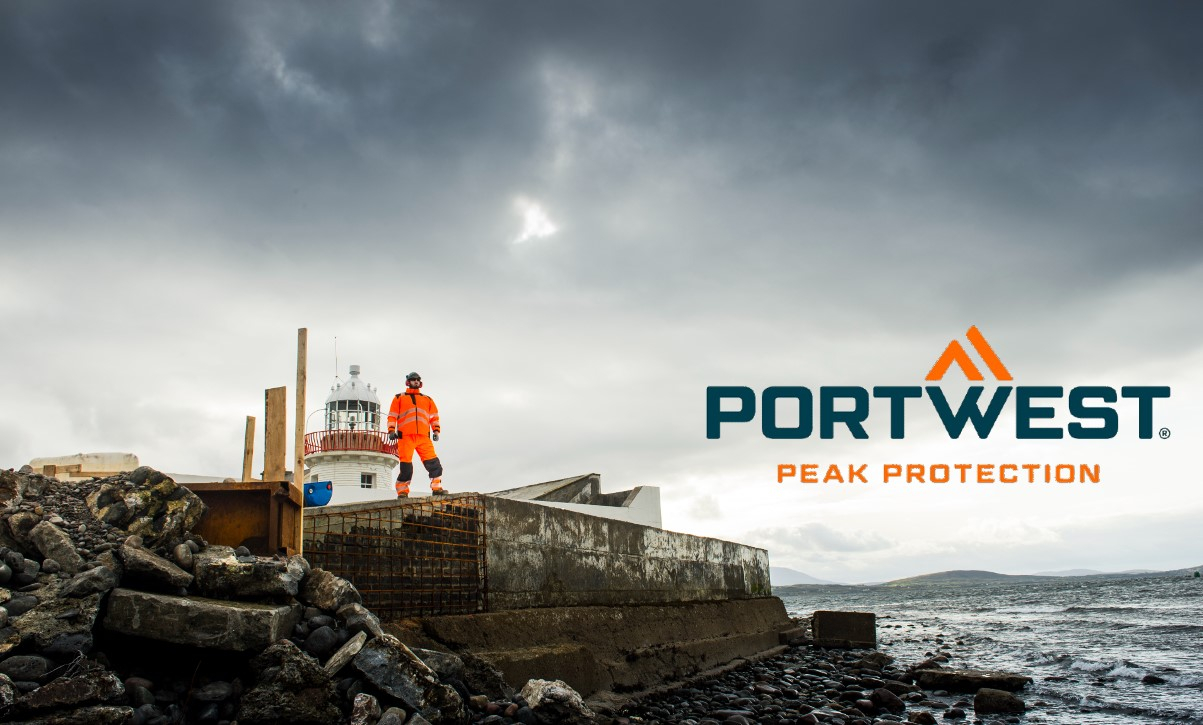 Un lavoratore che indossa indumenti arancioni ad alta visibilità si trova su un cantiere roccioso accanto a un faro, con il logo Portwest e lo slogan "Peak Protection" nella foto. Esiste un collegamento che porta al nostro abbigliamento da lavoro multinorma.