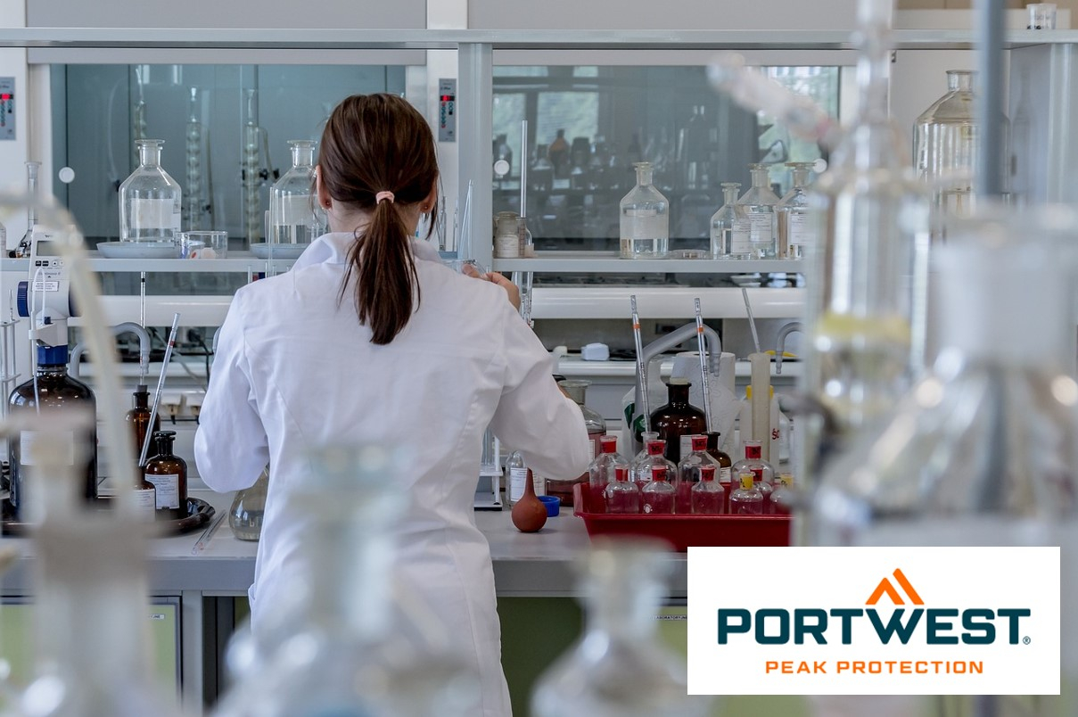 Una donna con i capelli castano scuro legati indietro indossa un camice da laboratorio bianco e lavora in un laboratorio moderno con varie attrezzature da laboratorio e bottiglie chimiche. In basso a destra nell'immagine c'è il logo "Portwest Peak Protection". C'è un collegamento che porta a tutti i nostri camici da laboratorio.