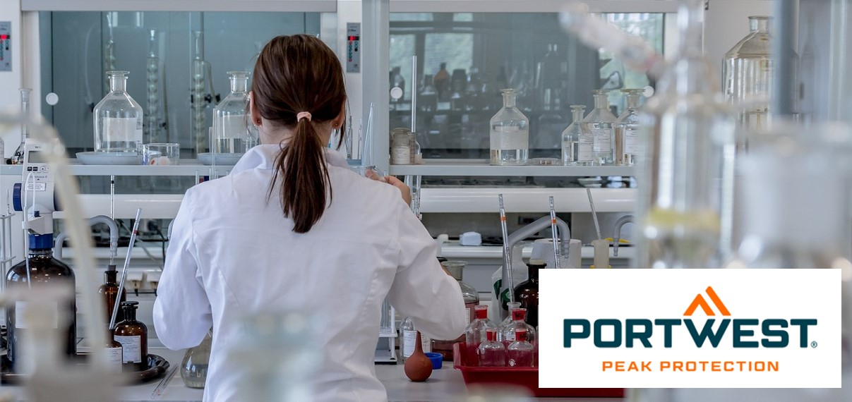 Una donna con i capelli castano scuro legati indietro indossa un camice da laboratorio bianco e lavora in un laboratorio moderno con varie attrezzature da laboratorio e bottiglie chimiche. In basso a destra nell'immagine c'è il logo "Portwest Peak Protection".