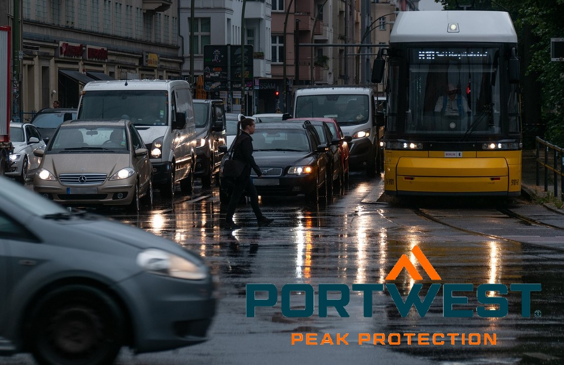 Scena di strada sotto la pioggia. Numerose auto e autobus sono parcheggiati agli incroci con i fari accesi. Il logo dell'azienda Portwest in arancione e blu si trova nell'angolo in basso a destra dell'immagine. C'è un collegamento che porta alle nostre giacche antipioggia.