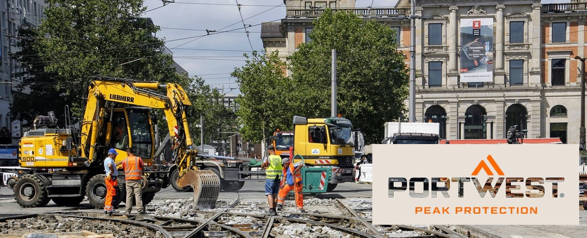 Operai edili in indumenti di sicurezza arancioni lavorano in un cantiere edile in città. Sullo sfondo si possono vedere escavatori, camion e un edificio storico. Il logo Portwest e lo slogan "Peak Protection" sono mostrati nella parte inferiore dell'immagine.