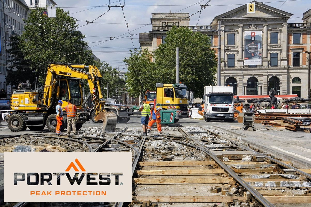 Operai edili in indumenti di sicurezza arancioni lavorano in un cantiere edile in città. Sullo sfondo si possono vedere escavatori, camion e un edificio storico. Il logo Portwest e lo slogan "Peak Protection" sono mostrati nella parte inferiore dell'immagine. È presente un collegamento che porta alla pagina dell'articolo sul tappo di raffreddamento.